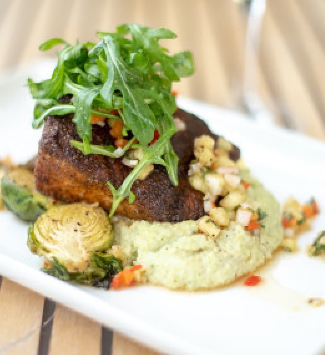 A plated dish featuring a blackened fish fillet topped with arugula, served over a bed of seasoned vegetables and a creamy green sauce, presented on a white plate.